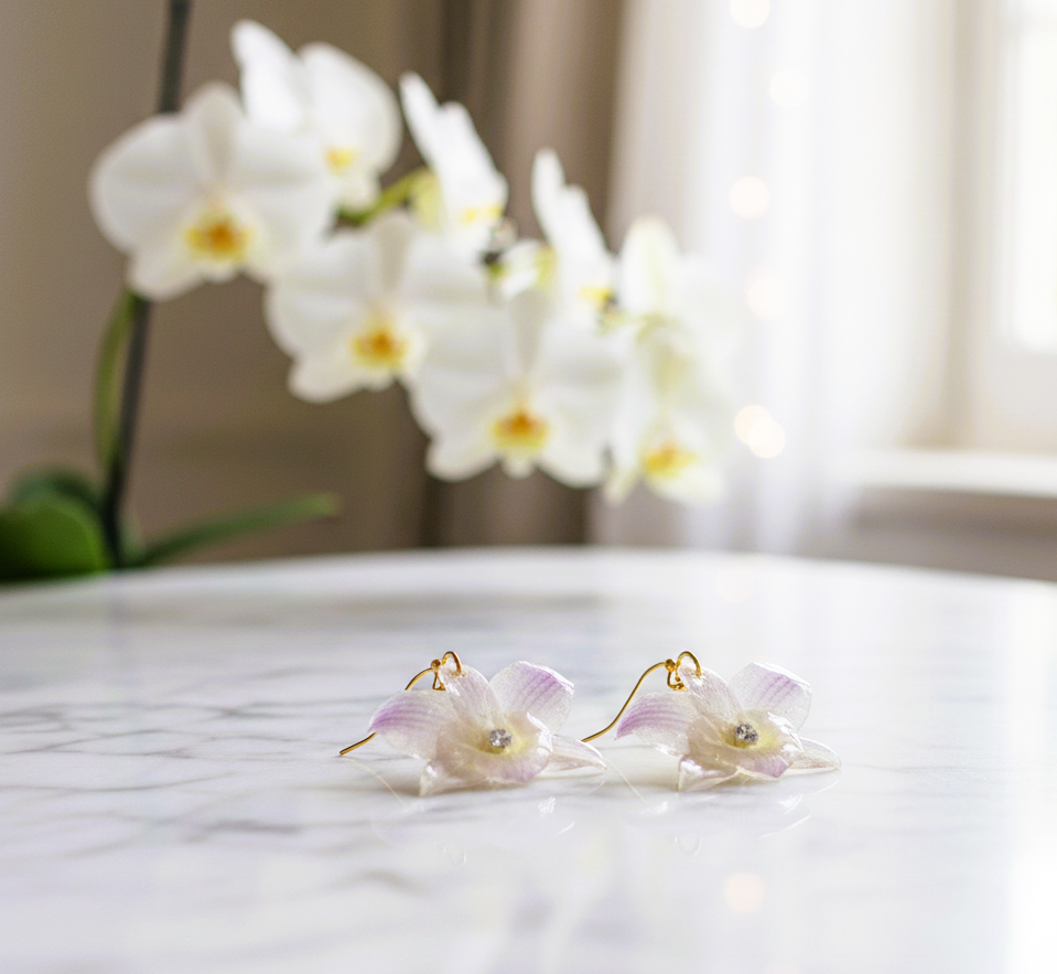 L'eleganza della natura da indossare: orchidee Dendrobium vere, eteree e senza tempo. Primo piano di orecchini pendenti realizzati con vere orchidee Dendrobium bianche e sfumature lilla preservate in resina trasparente, con dettaglio di cristallo centrale e ganci dorati.