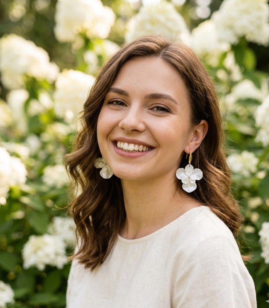 La luminosità degli Hydra White Drop: orecchini con fiori veri per un look fresco e naturale. Primo piano di una ragazza sorridente che indossa orecchini pendenti a forma di ortensia bianca con gancio dorato, sfondo con fiori di ortensia bianchi sfocati.
