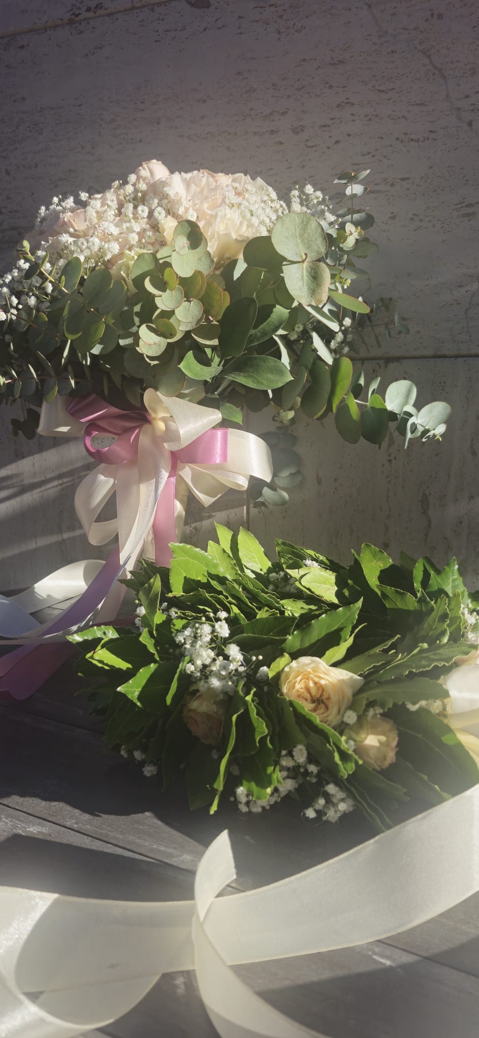 Kit proclamazione laurea composto da una corona di alloro con rose crema e nebbiolina e un bouquet di fiori freschi nei toni del rosa e eucalipto con fiocchi di raso, presentato su un piano di legno.