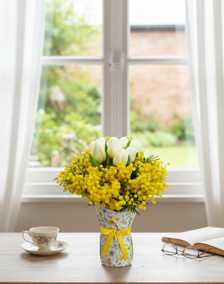 Bouquet di mimosa gialla e tulipani bianchi in confezione regalo floreale su un tavolo in legno davanti a una finestra.