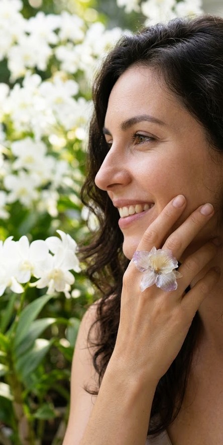 Primo piano di una donna che indossa un anello artigianale realizzato con un'orchidea Dendrobium vera stabilizzata in resina trasparente, sfondo con orchidee bianche sfocate.